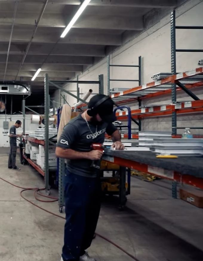 A Cryptic Air fabricator assembling linear slot diffusers on the shop floor, finished product stacked along the bench beside pallet racking