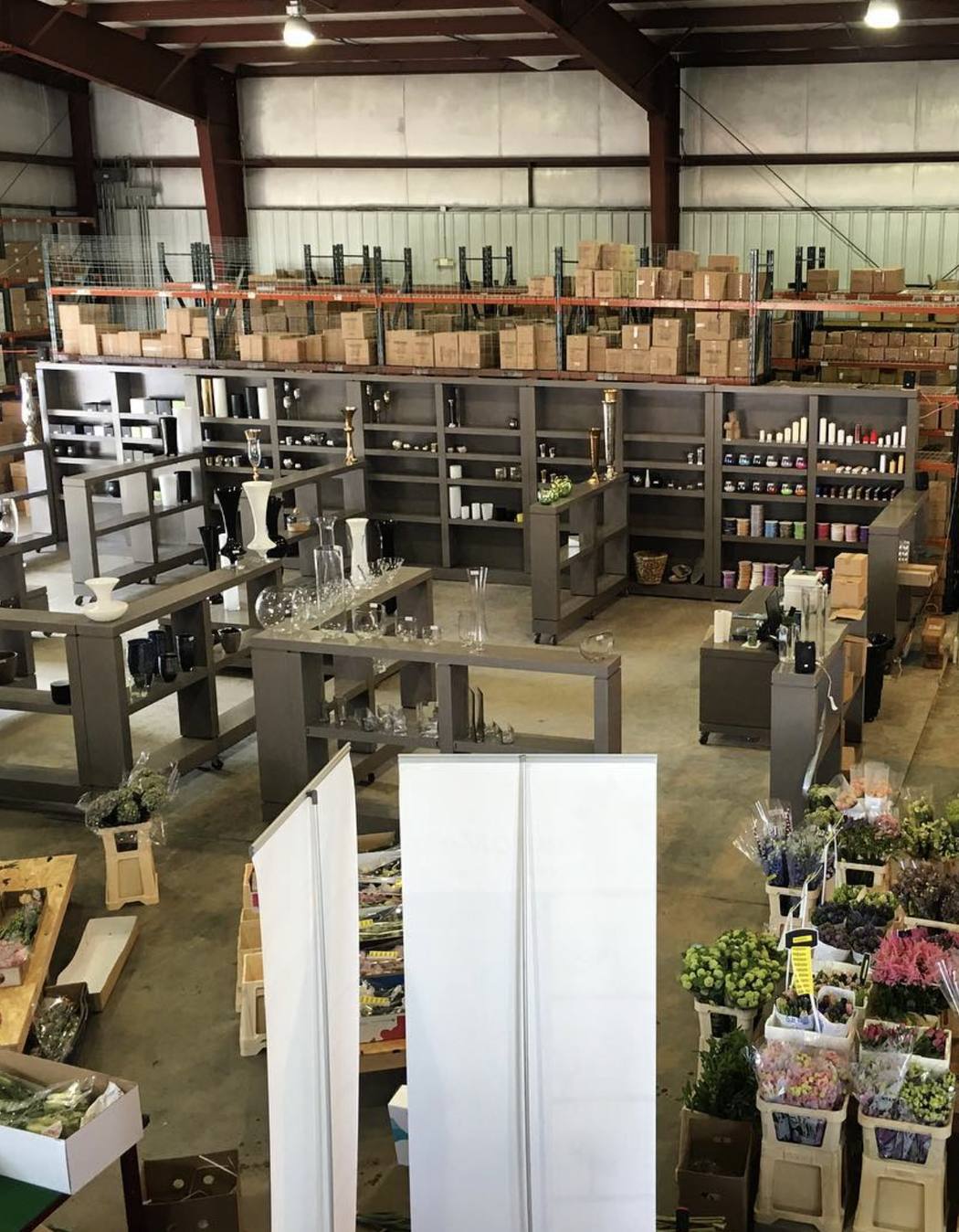Display shelving of vases, candleholders and ribbon in front of pallet racking, with fresh cut stems in buckets on the warehouse floor